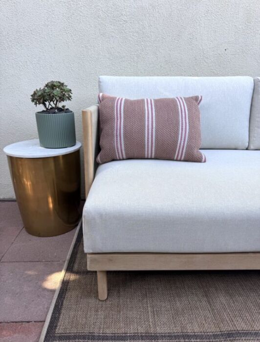 A modern outdoor sofa with a striped pillow sits next to a round gold side table holding a potted succulent, against a light-colored wall.