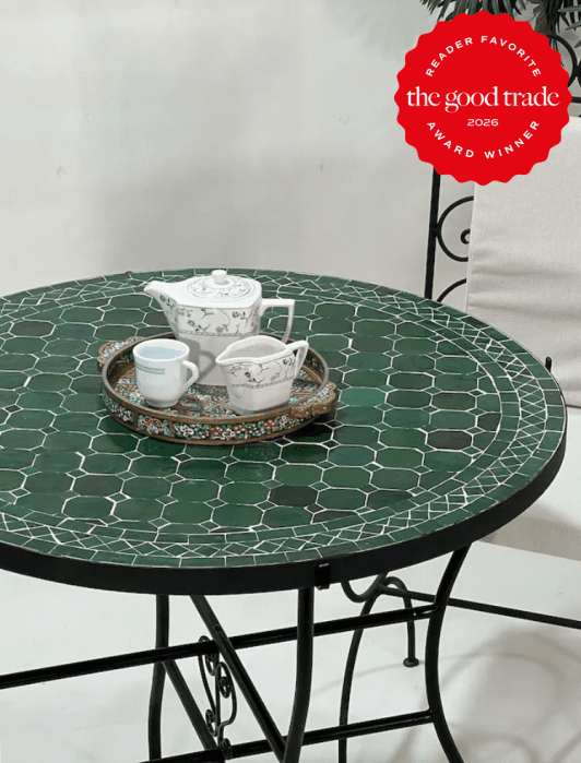 A round green mosaic table with a tea set on a tray; includes a teapot, cup, and small bowl. A red award badge from The Good Trade is in the upper right corner.