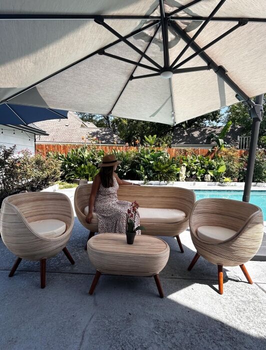 A woman in a patterned dress and hat sits on an outdoor sofa under a large umbrella, surrounded by matching chairs and a table, next to a swimming pool with greenery in the background.
