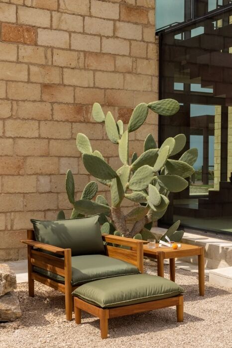 A wooden lounge chair with green cushions and a matching table sit on gravel beside a stone wall, near a large prickly pear cactus.