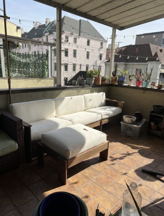 A sunlit balcony with tiled floor, a white cushioned wooden sofa and ottoman, potted plants on the railing, and various household items in the corners.
