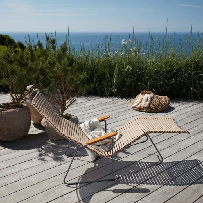 A lounge chair with wooden slats and metal frame sits on a deck overlooking the ocean, surrounded by potted plants and a woven basket.