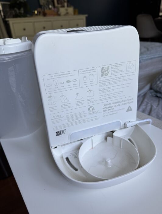 A white portable humidifier with its water tank removed, showing the open base and instruction labels on the inside panel. The unit sits on a white surface in a bedroom.
