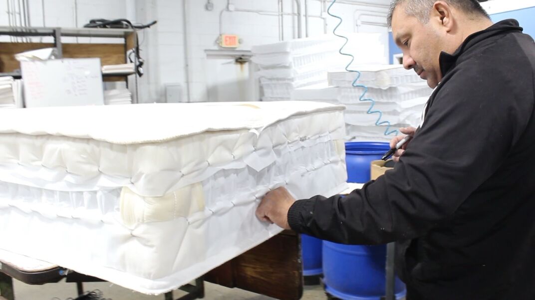 A man inspects or assembles a mattress in a manufacturing facility, using tools with blue barrels and stacked mattresses in the background.