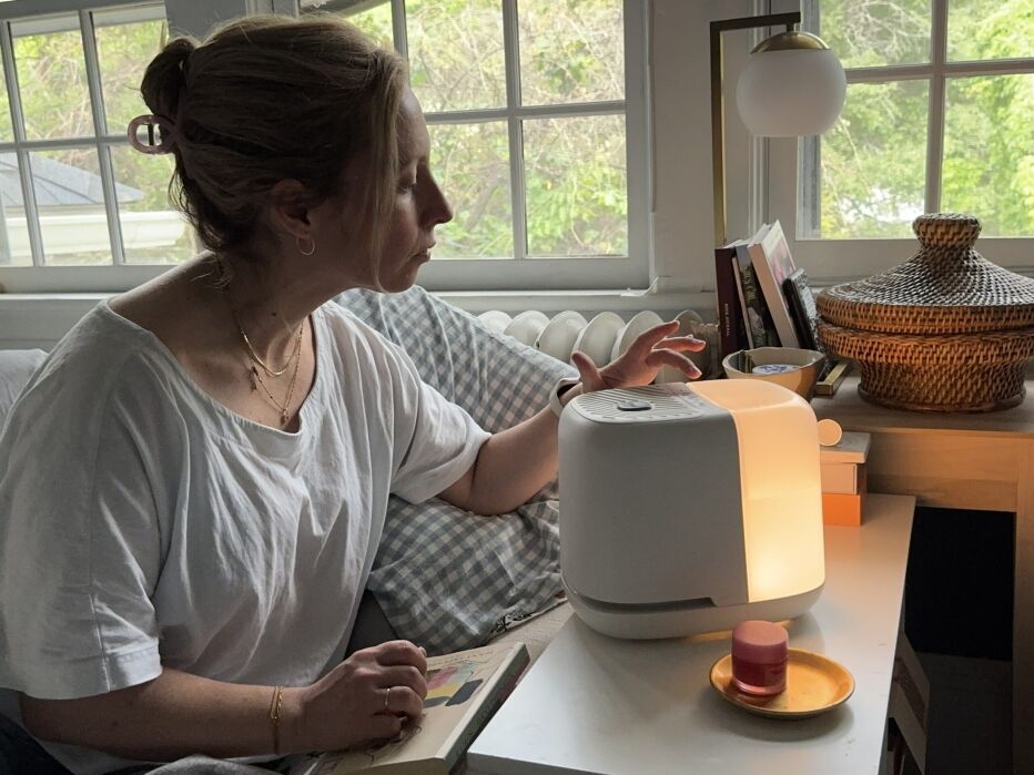 A woman sits on a couch next to a table, reaching toward a white humidifier or diffuser that is lit, with a small container and a book nearby.