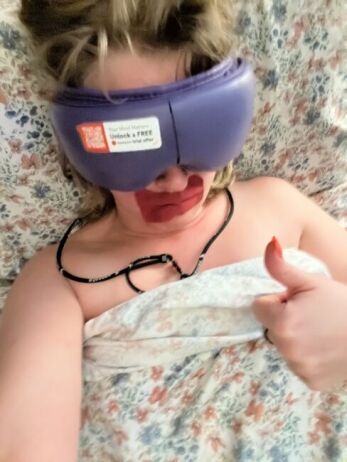 A person lying on a bed with a purple sleep mask and red substance around their mouth gives a thumbs up while partially covered by a floral blanket.