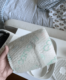 A hand holds a used and dirty air purifier filter above a white table with purifier parts; a bed with patterned sheets is in the background.
