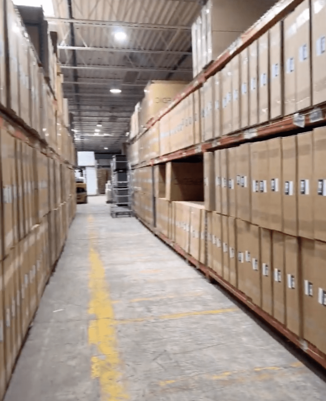Long aisle in a warehouse lined with tall shelves filled with large cardboard boxes on both sides; yellow line marks run along the concrete floor.