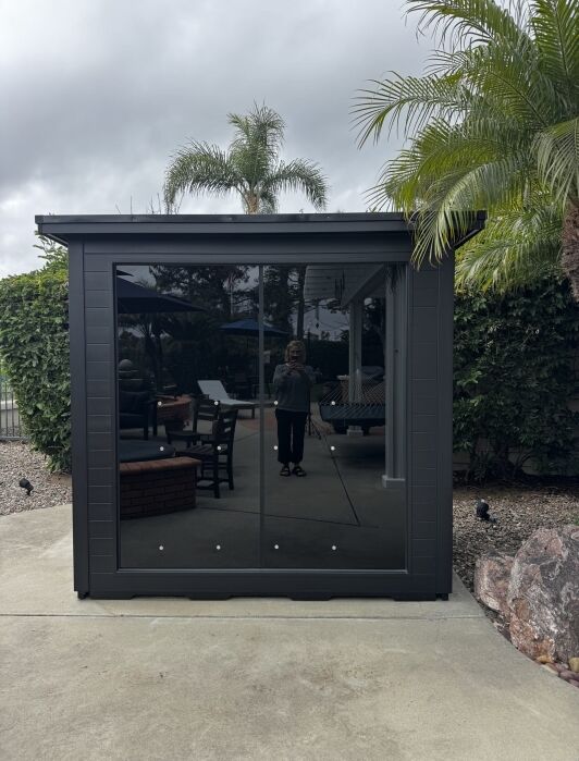 A person is reflected in the glass doors of a modern outdoor shed, surrounded by patio furniture, palm trees, and landscaping under a cloudy sky.