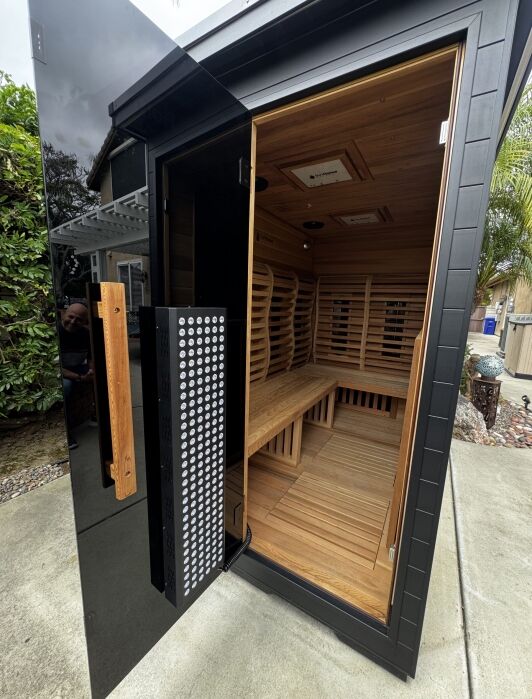A modern outdoor infrared sauna with a glass door, wooden interior benches, and infrared panels, situated on a concrete patio near plants and trees.