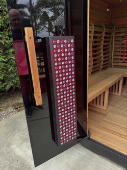 A red light therapy panel is mounted on the door of a wooden sauna with benches inside; a person stands outside the open door.
