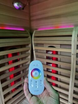 A hand holds a remote control with a color wheel inside a wooden sauna with red infrared panels and purple LED lights on the ceiling.