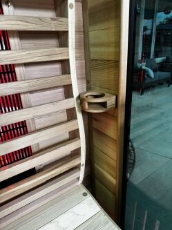 Wooden bench and slatted backrest inside a sauna, with a small wooden cup holder attached to the wall, and glass door showing a view outside.