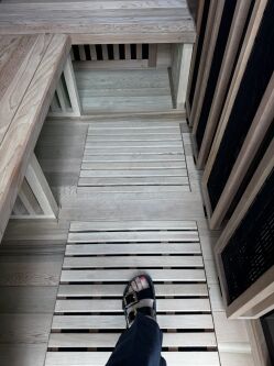 A person wearing black sandals stands on a wooden slatted floor inside a light-colored wooden sauna or steam room.