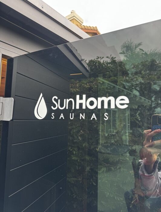 Glass door with "Sun Home Saunas" logo and reflection of person taking the photo, with plants and building visible in the background.