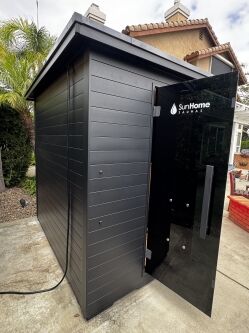 A black outdoor sauna with a glass door labeled "SunHome Saunas" stands on a concrete patio near a house and garden.