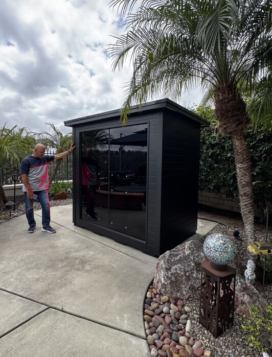 A man stands next to a small black outdoor shed with glass doors, surrounded by palm trees, rocks, and garden decorations on a concrete patio.