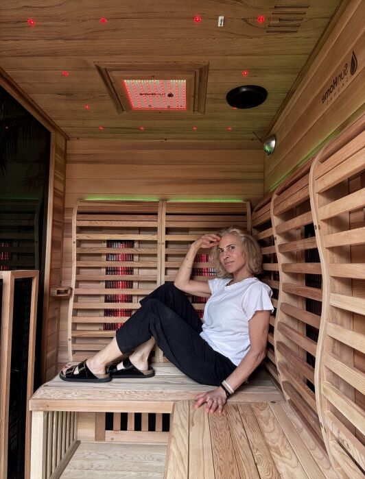A woman sits on a wooden bench inside a modern infrared sauna, wearing a white t-shirt, black pants, and sandals, with colored LED lights on the ceiling.
