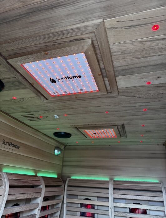 Wood-paneled interior of a sauna with SunHome infrared panels on the ceiling, small red LED lights, and green accent lighting along the walls.