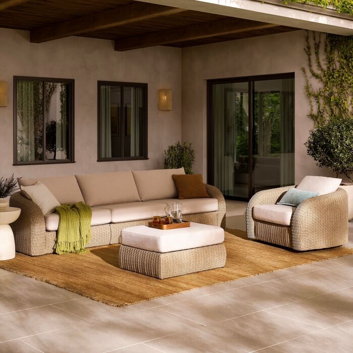 Outdoor patio with wicker furniture, including a sofa, armchair, and ottoman on a tan rug, with green plants and neutral-colored walls in the background.