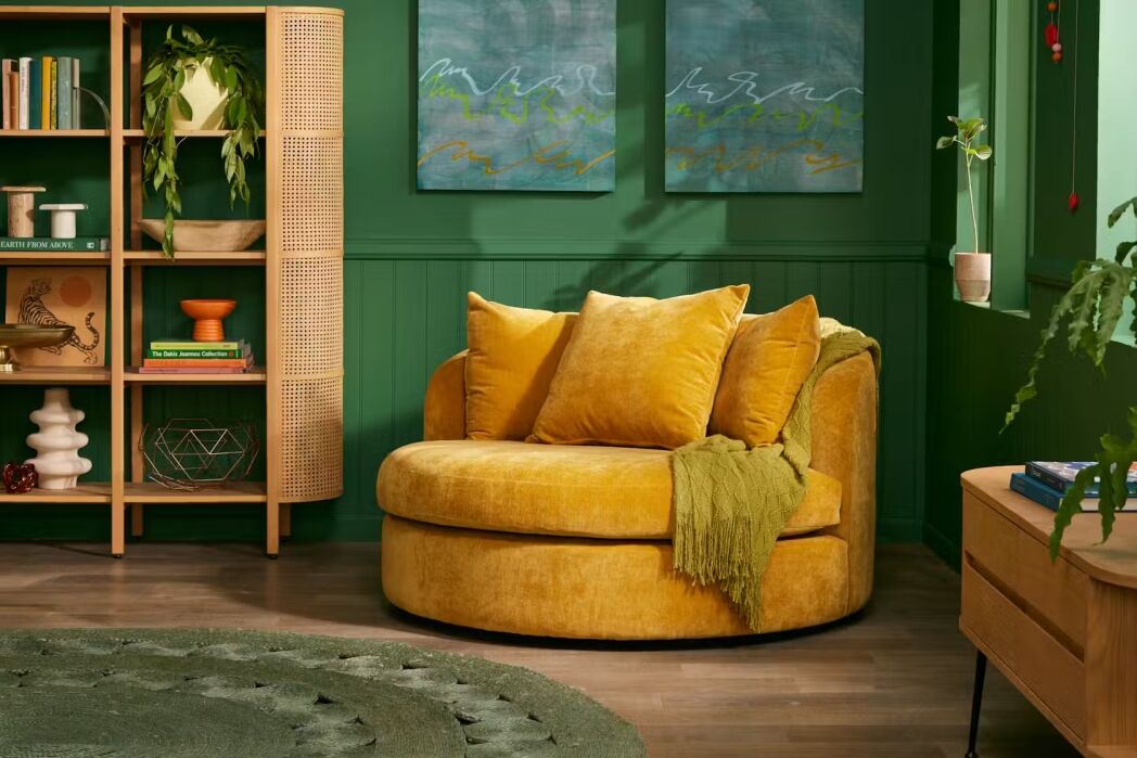 A cozy yellow velvet loveseat with green throw blanket in a green-themed living room, featuring plants, a bookshelf, wall art, and a round rug.