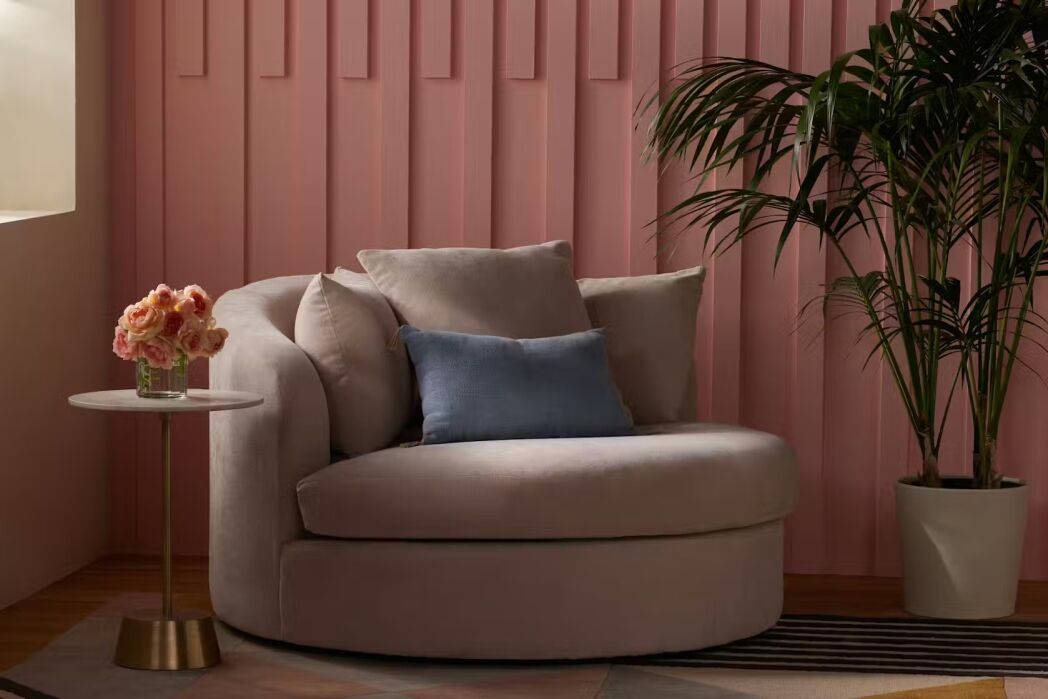 A large round beige chair with a blue pillow stands next to a small round table with pink flowers; a potted plant is nearby, against a pink paneled wall.