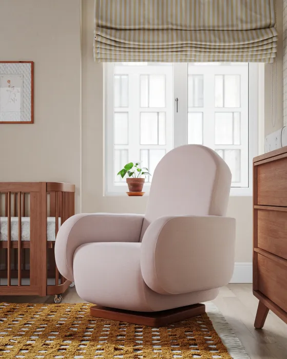 A modern, light pink armchair sits on a patterned rug in a nursery with a wooden crib, chest of drawers, and a potted plant by a window with striped Roman shades.