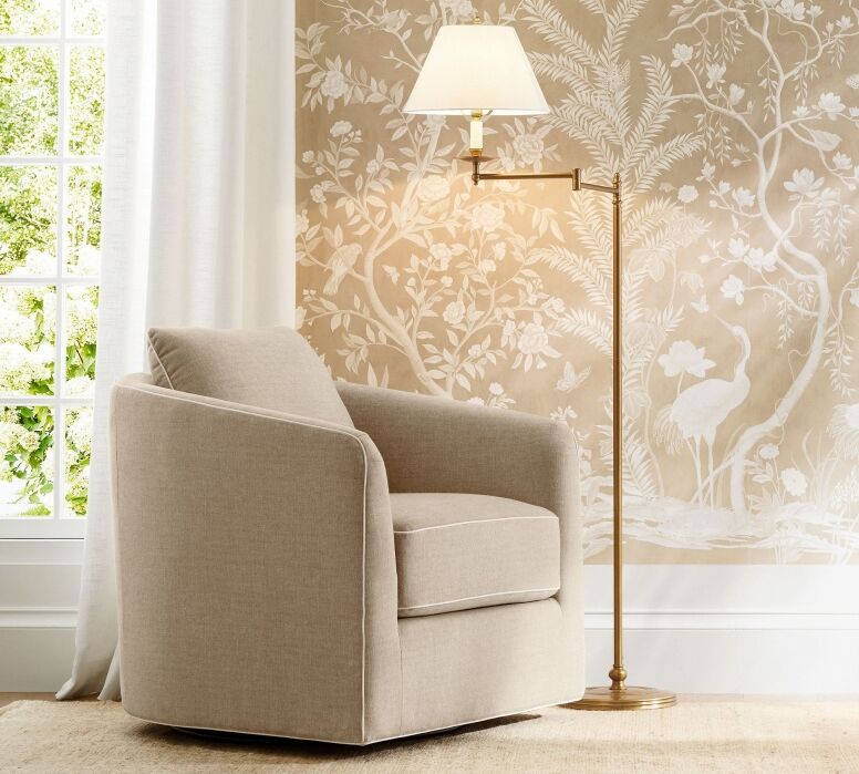 A beige armchair sits next to a brass floor lamp in front of a window and a wall with a light-colored nature-themed mural.