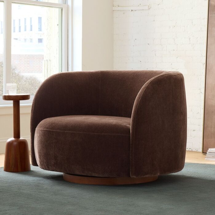 A brown upholstered swivel chair with a curved back sits next to a small wooden side table in a bright room with white brick walls and large windows.