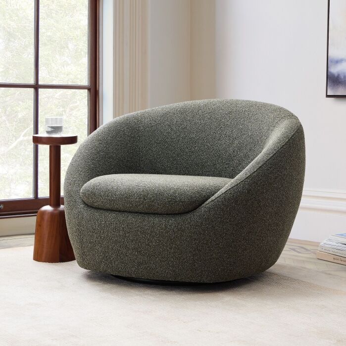 A modern, rounded gray armchair sits on a light rug next to a small wooden side table with a glass, in a bright room with large windows.