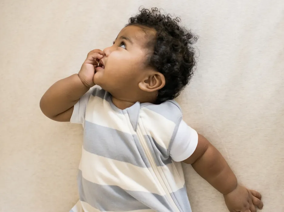 Baby in a striped sleep sack lying on a light-colored surface, looking upward and sucking thumb.