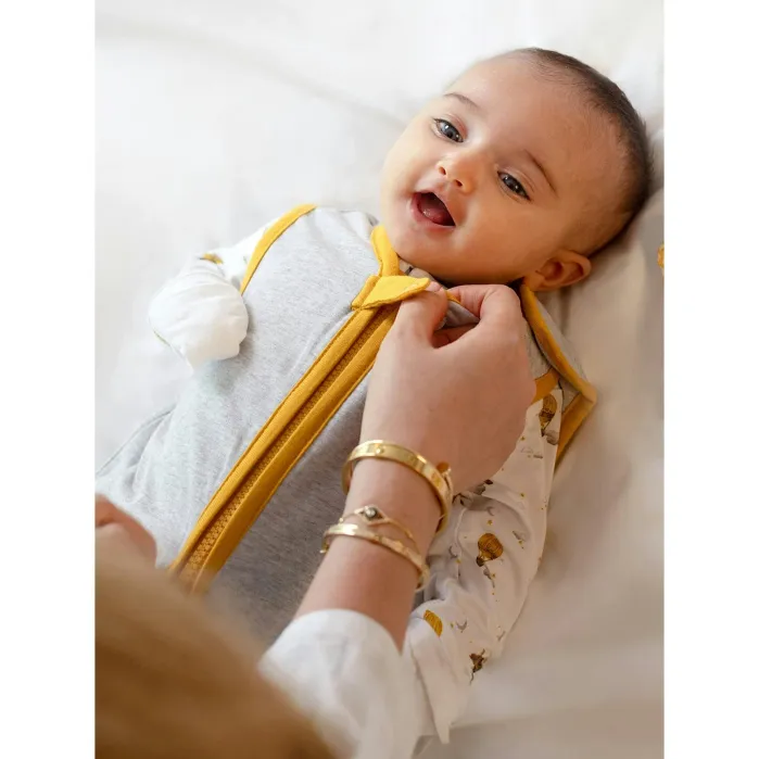 An adult fastens a yellow and gray sleeping bag on a baby lying on a white surface.