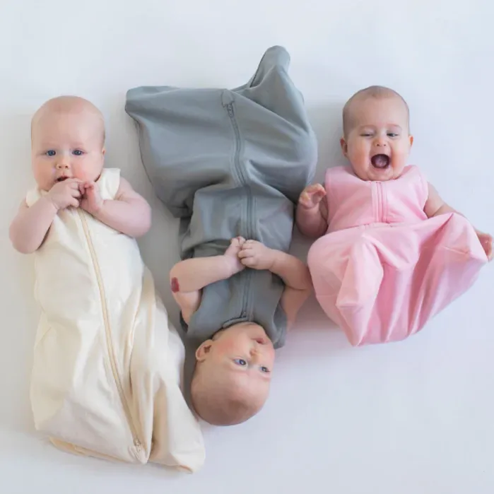 Three babies are lying on a white surface, each wearing a different colored sleep sack: cream, gray, and pink. The baby in the middle is upside down.