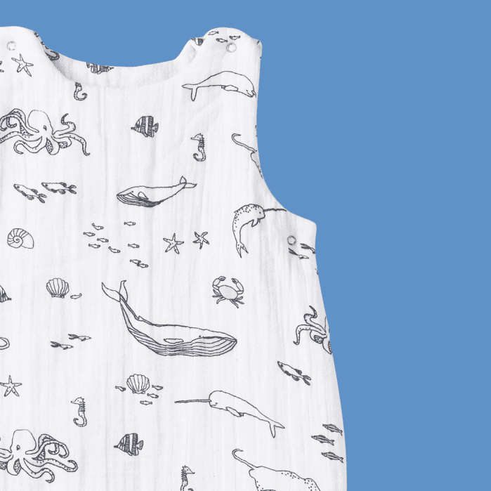 White sleeveless baby romper with black line drawings of sea creatures, including whales, octopus, crabs, and shells, displayed against a solid blue background.