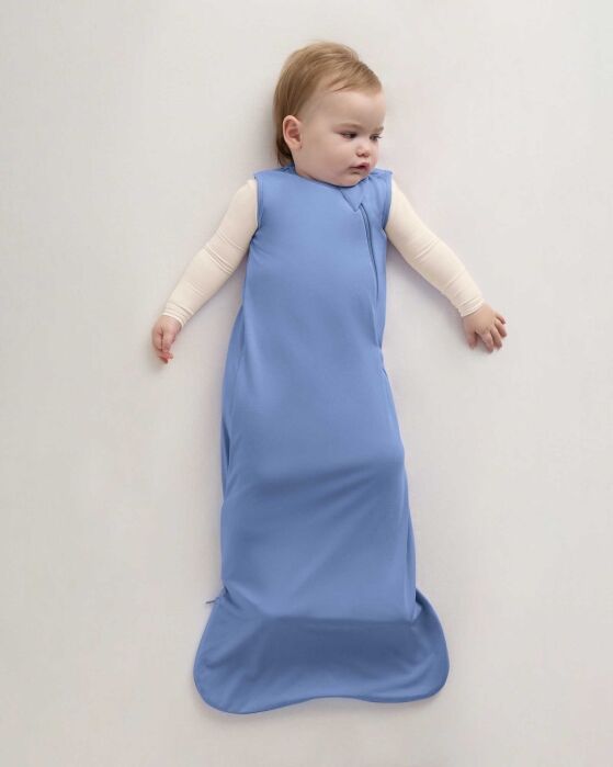 A baby lies on a light surface wearing a long blue sleeveless sleep sack over a cream long-sleeve top.