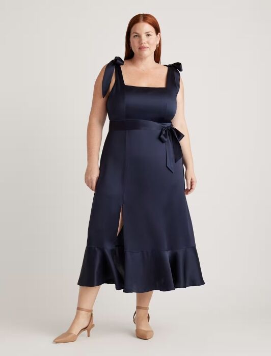 A woman stands wearing a navy blue sleeveless midi dress with bow straps, a waist tie, a front slit, and a ruffled hem, paired with nude low-heel shoes.