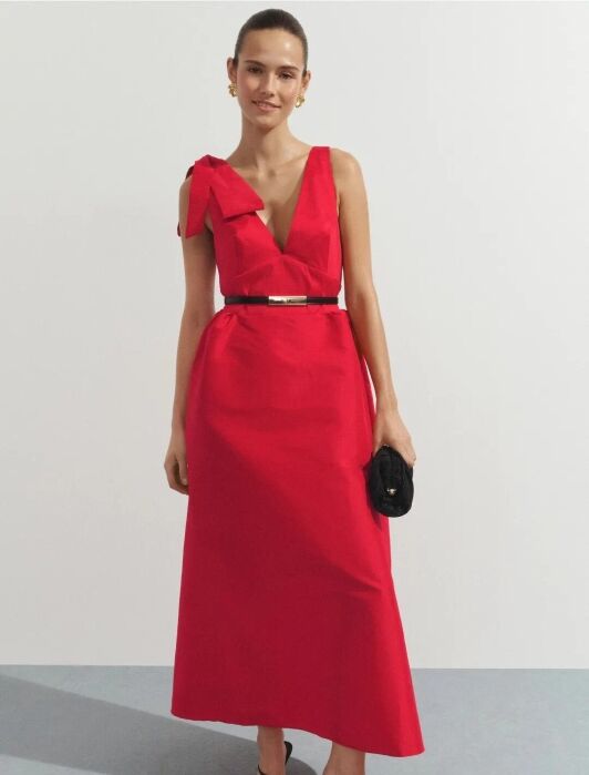 A woman stands wearing a sleeveless, ankle-length red dress with a deep V-neck, a bow on one shoulder, black belt, black sandals, and holding a black clutch.