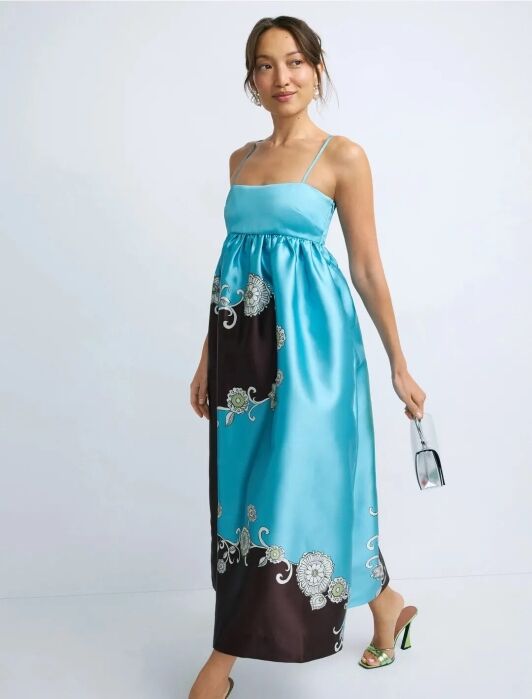Woman wearing a sleeveless blue and brown maxi dress with floral embroidery, holding a small clear handbag, and wearing green heeled sandals.