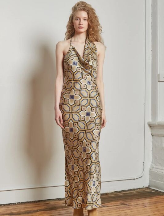 A woman stands indoors wearing a sleeveless, floor-length dress with a geometric gold, blue, and tan pattern, paired with tan shoes.