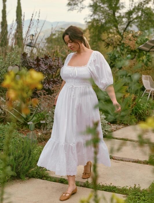 A woman in a white, long, puff-sleeve dress walks on a stone path through a lush garden, surrounded by greenery and flowers.