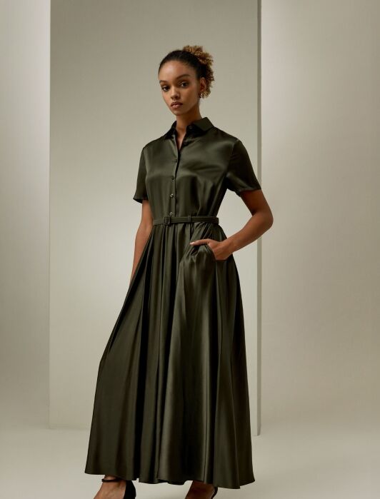 A woman stands in a studio wearing a dark green, short-sleeve, button-up, belted maxi dress with pockets and black heeled sandals.
