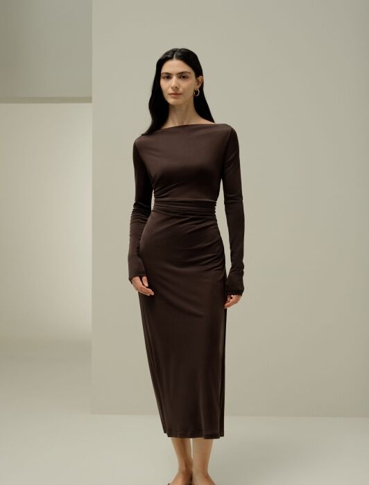 A woman stands indoors wearing a long-sleeved, dark brown, ankle-length dress and brown flats, against a plain light background.