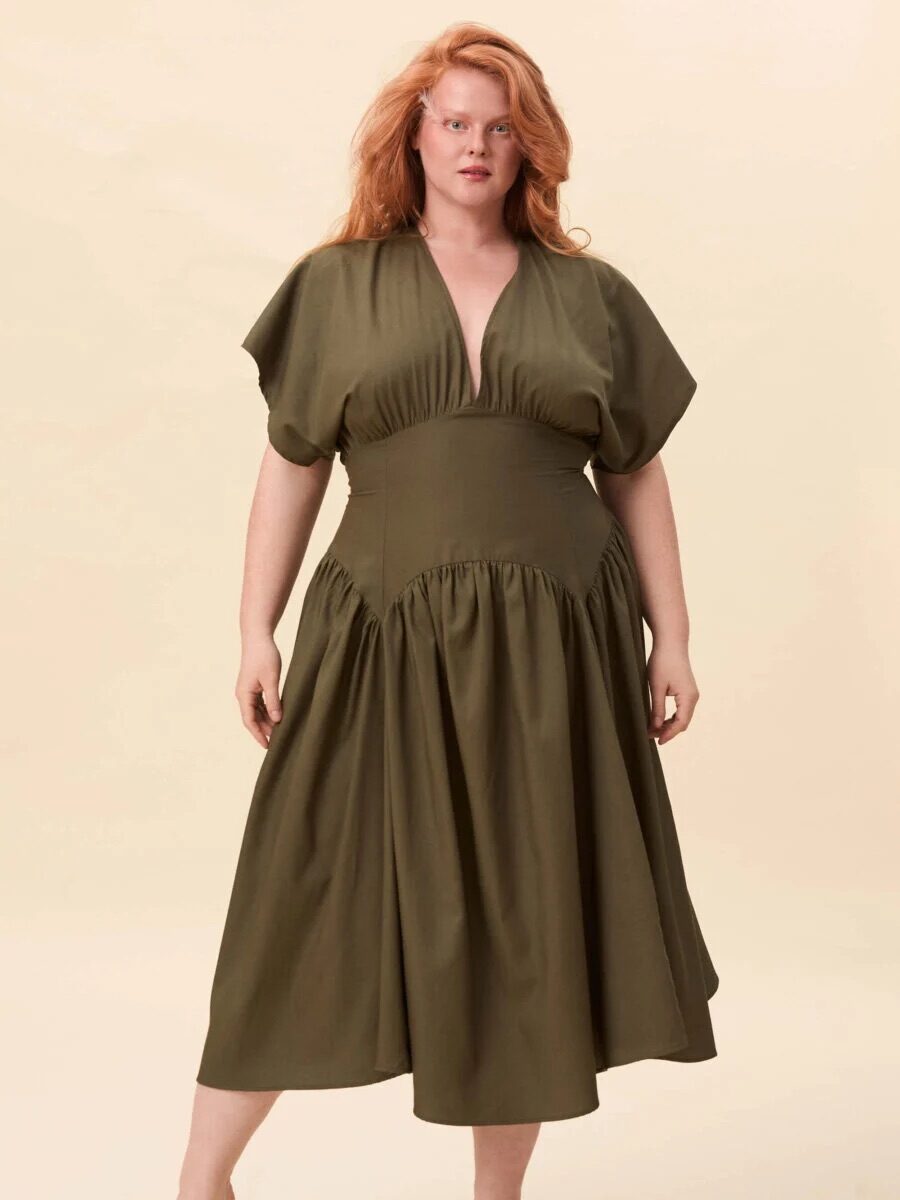 A person wearing a green, short-sleeved, v-neck midi dress with a fitted waist and full skirt, standing on a neutral background.