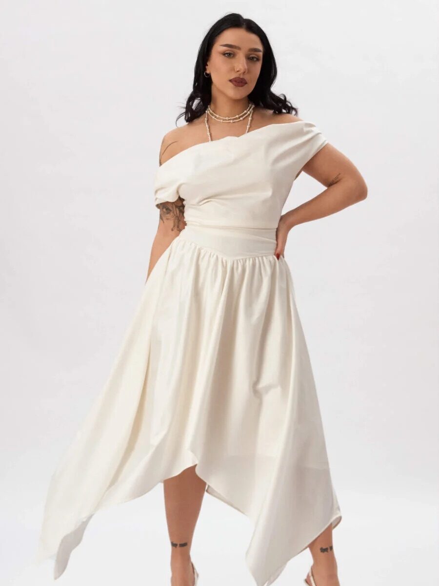 Woman wearing an off-shoulder white asymmetrical dress, white high heels, and layered necklaces, standing against a plain white background.
