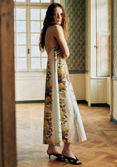 A woman with long wavy hair wears a sleeveless, backless floral dress and black heels, standing in a sunlit room with wooden floors and patterned wallpaper.
