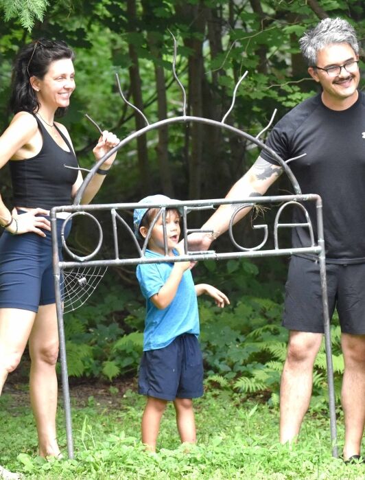 A woman, man, and two children stand outdoors next to a metal sign reading "OMEGA," surrounded by green foliage.