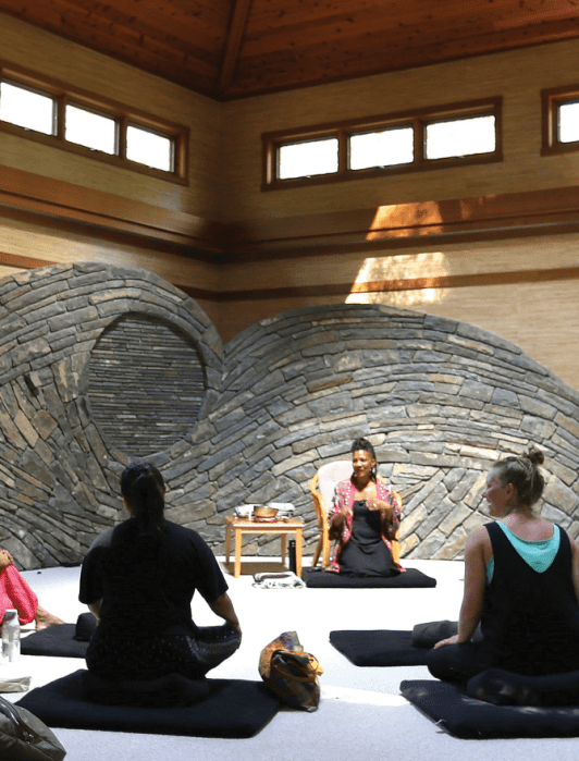 A group of people sit on cushions in a serene room with wooden walls and a stone wave feature, listening to a woman speaking at the front.