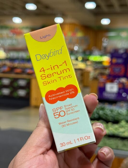 A hand holds a box of Daybird 4-in-1 Serum Skin Tint SPF 50 in a grocery store produce section.