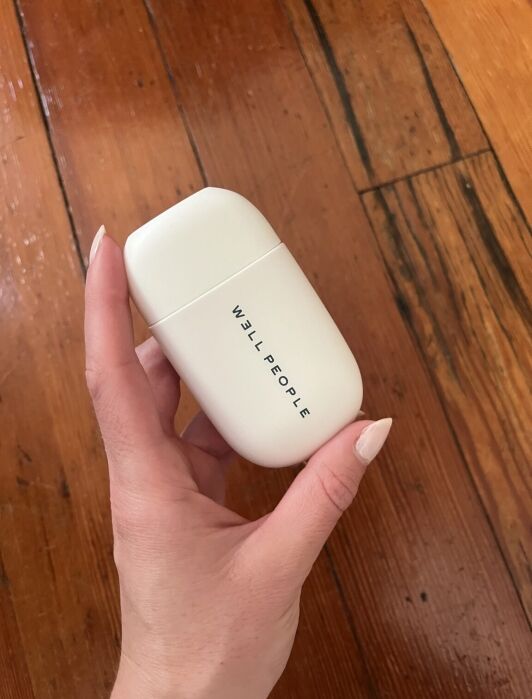 A hand holding a white, rectangular container labeled "WELL PEOPLE" against a wood floor background.