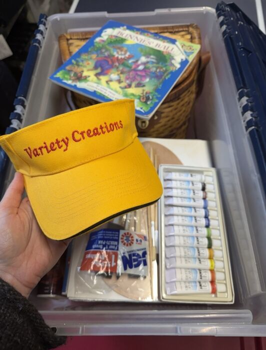 A hand holds a yellow visor reading "Variety Creations" above a storage bin containing paints, brushes, art supplies, a basket, and a children’s book.